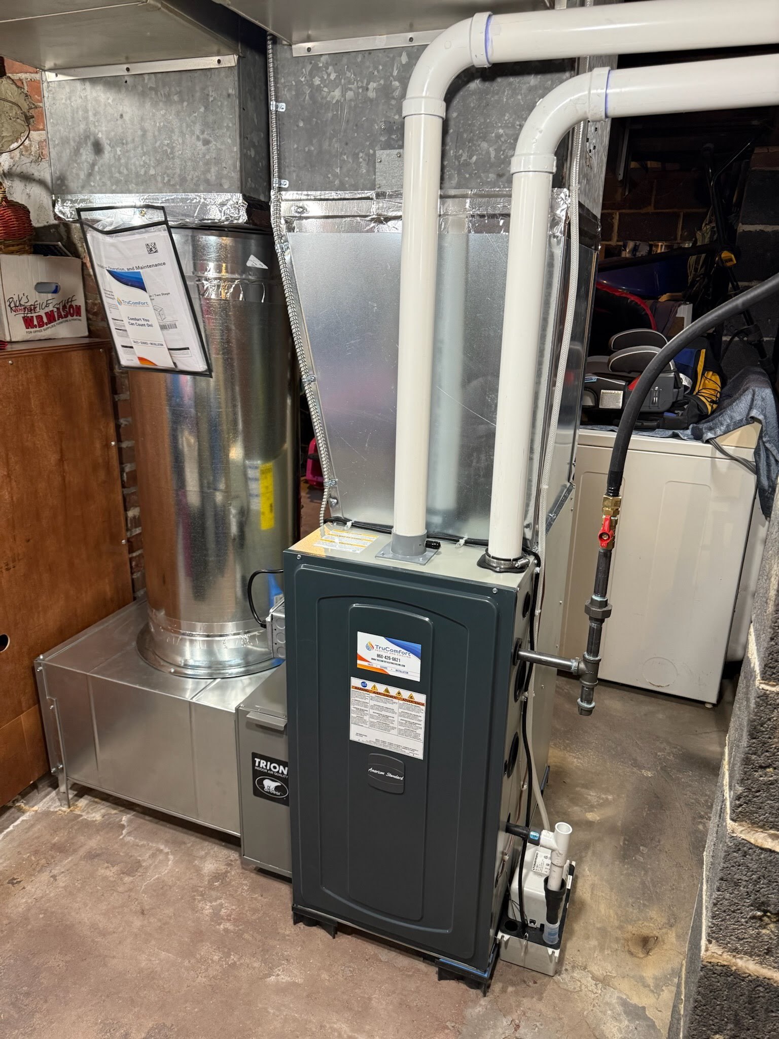 1c5ffab8e9eb2a487d54450e9842fdf2be43d69d-1 A residential HVAC unit neatly installed in a basement, featuring metal ductwork, PVC exhaust and intake pipes, a condensate pump, and electrical wiring. The equipment includes clearly labeled maintenance documentation. The surrounding basement area has concrete floors and storage items.